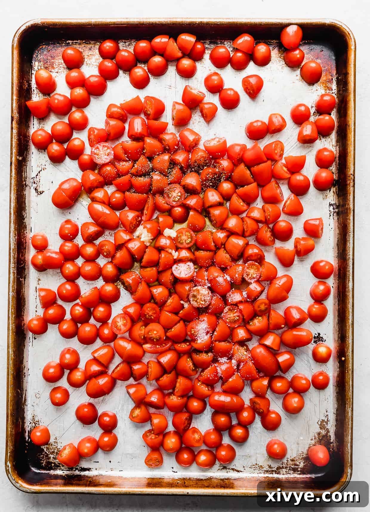 Small red tomatoes on a baking sheet topped with salt, pepper, and fresh garlic.