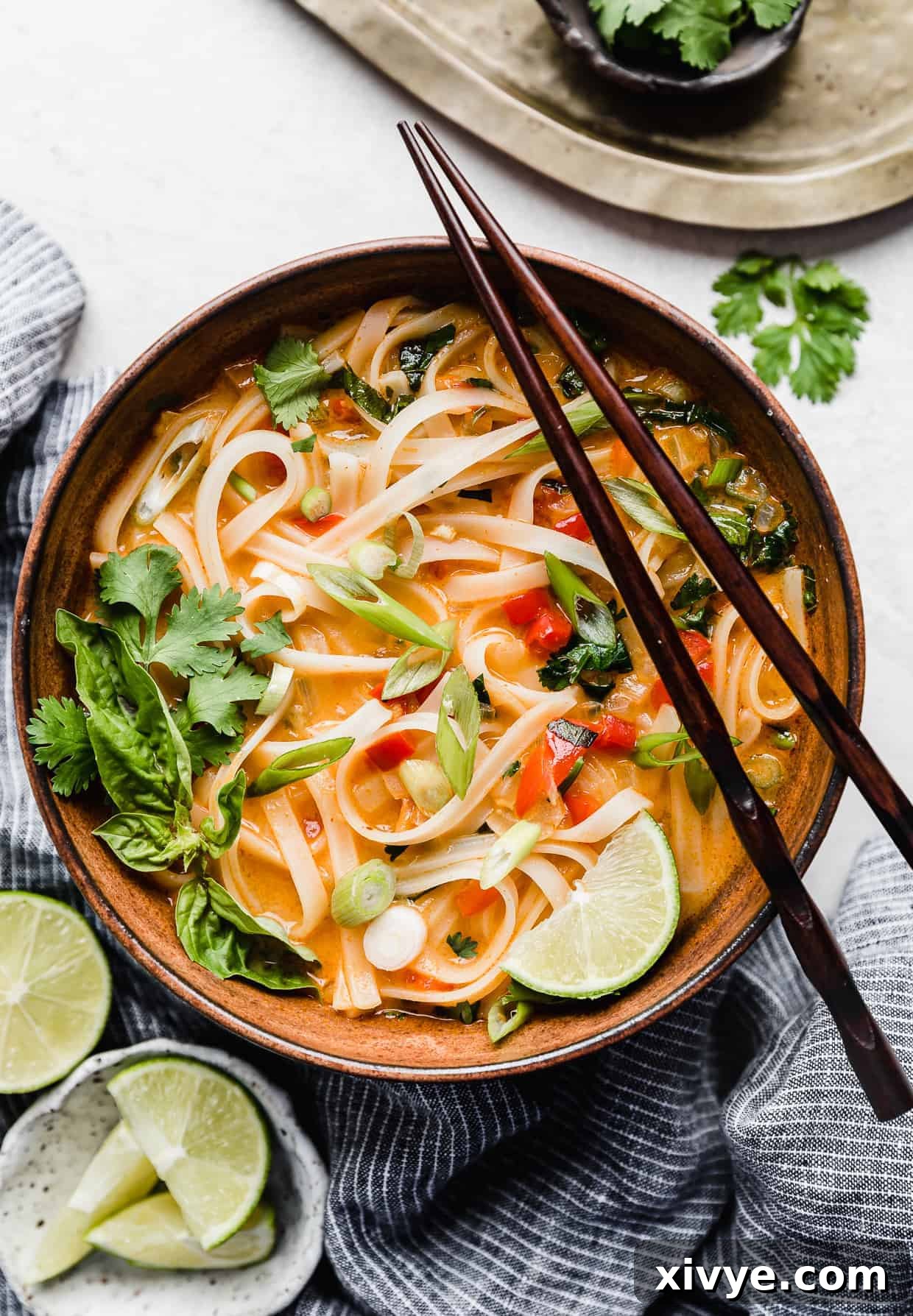 A steaming bowl of Thai Red Curry Noodle Soup, garnished with fresh herbs and lime wedges, with chopsticks resting on the rim.