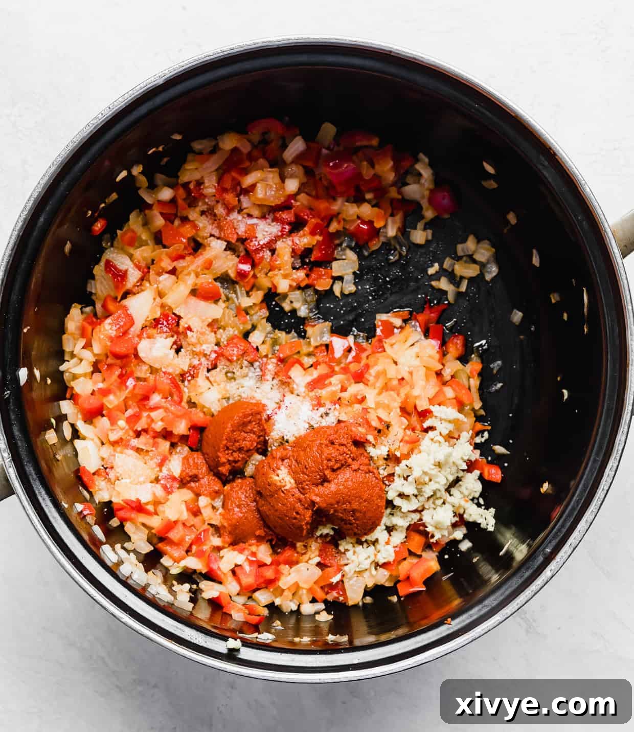 The same black pot, now containing cooked diced onion, red bell pepper, and red curry paste, stirred together and vibrant in color, preparing for the liquid ingredients of Thai Red Curry Noodle Soup.