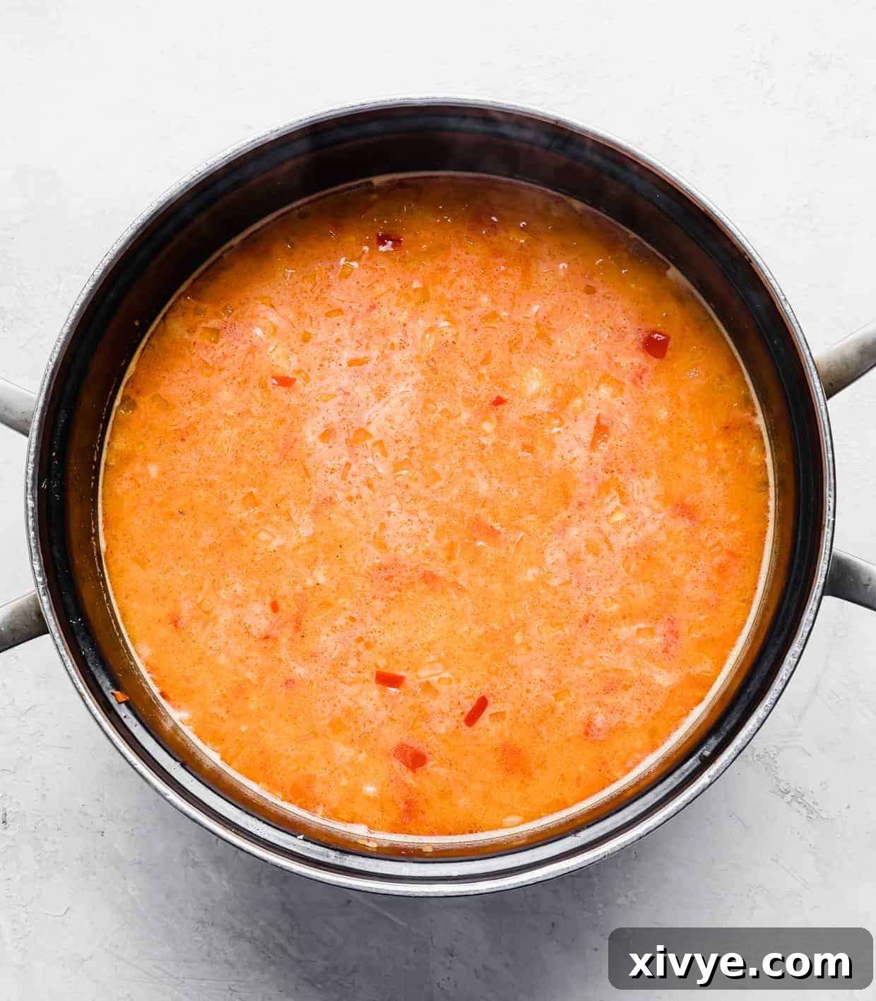 An overhead shot of a large black pot filled with a vibrant orange-colored Thai soup, now with broth and coconut milk added, simmering gently on a white background.