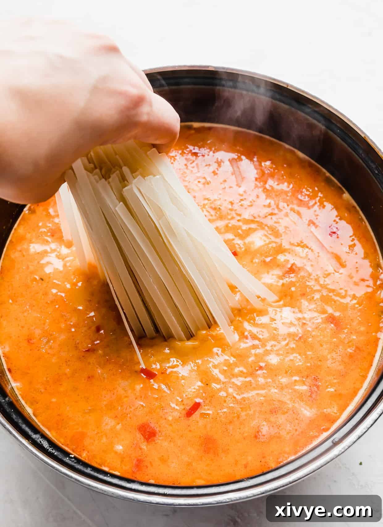 A hand gently placing linguine rice noodles into a simmering pot of Thai Red Curry Noodle Soup, ready to absorb the rich flavors.