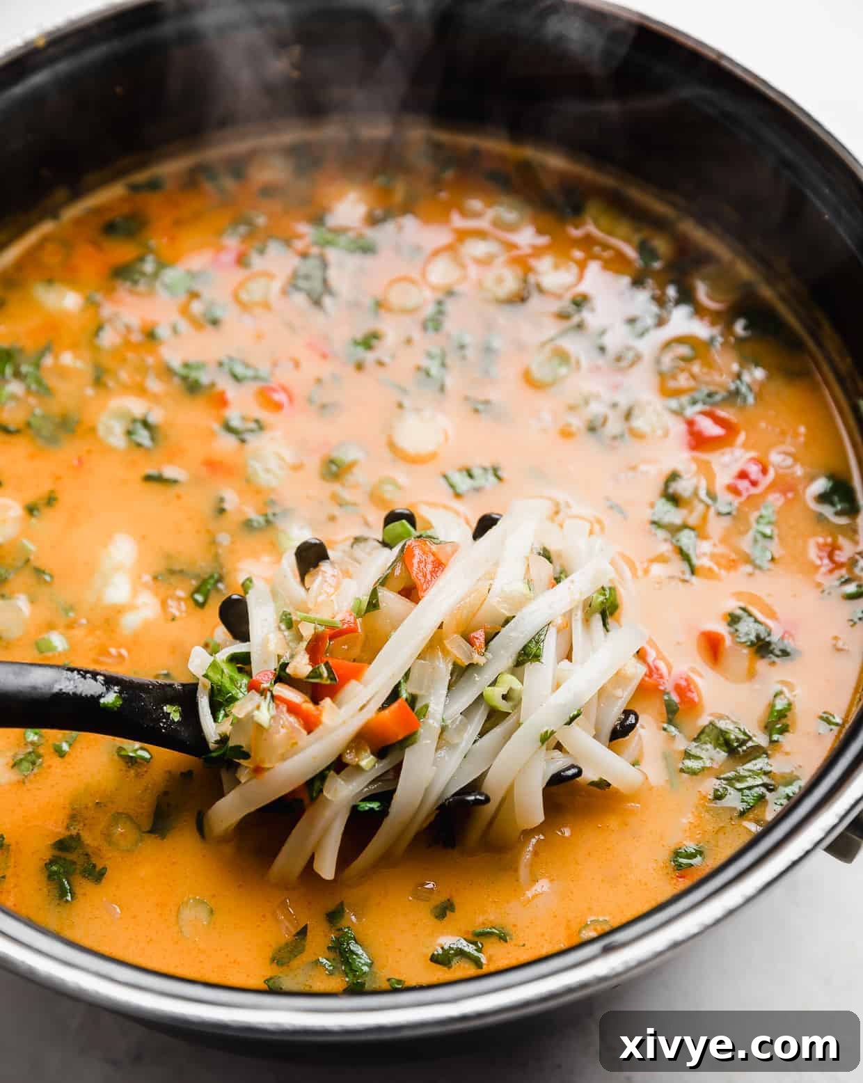 A spoon expertly scooping up a portion of linguine rice noodles along with rich broth from a large pot of Thai Red Curry Noodle Soup, showcasing its texture and abundance.