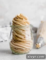Peanut butter frosting swirly piped into a small glass jar, with piping bag in background.
