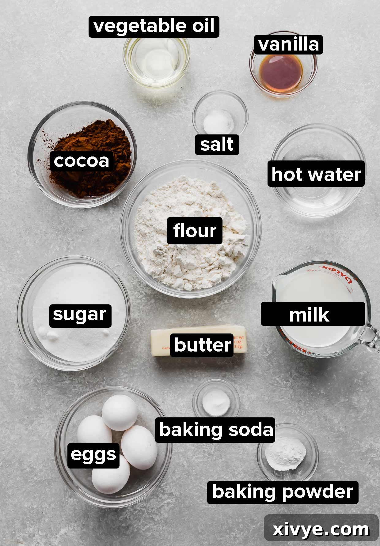 Ingredients used to make Chocolate Cake with Peanut Butter Frosting portioned into glass bowls on a light gray textured background.