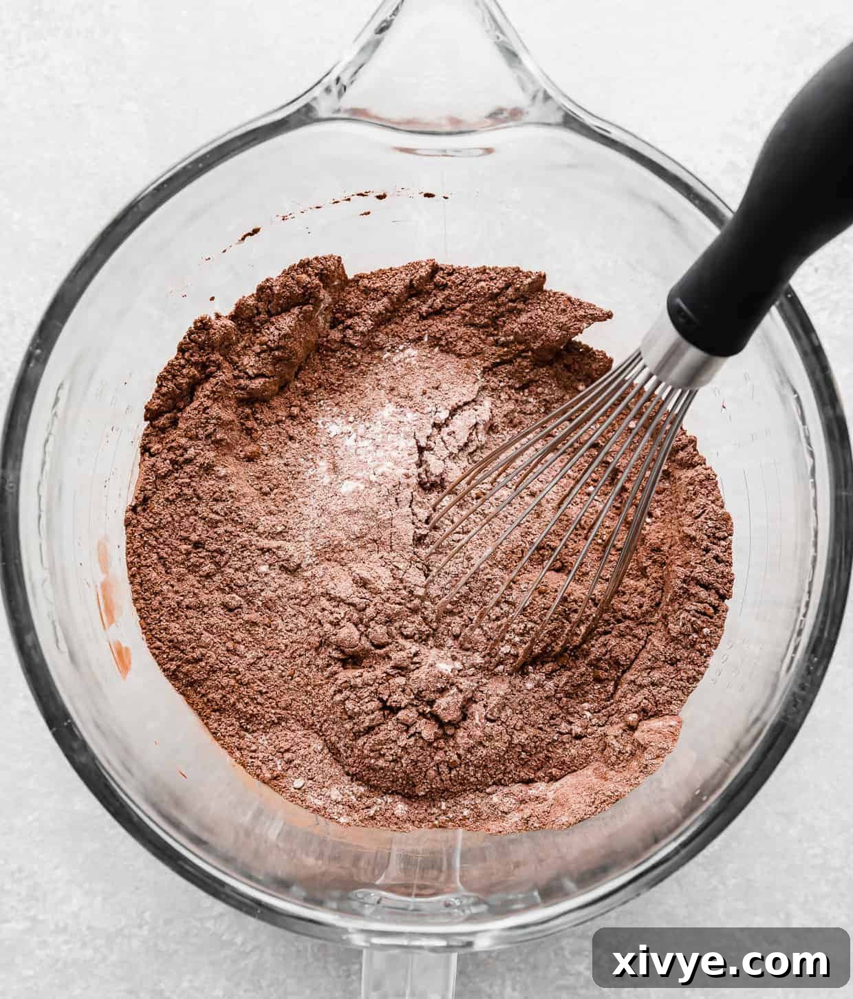 Dry ingredients for making chocolate cake in a glass stand mixer bowl, with a whisk stirring everything.