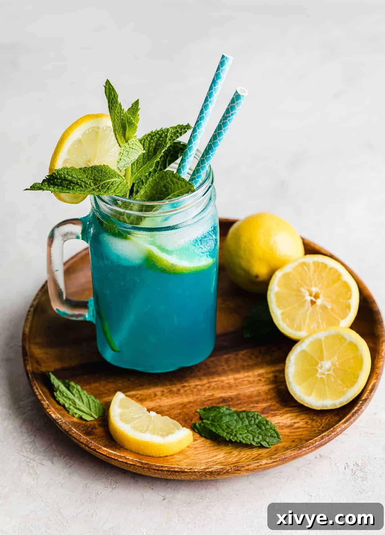 A sparkling glass of homemade Blue Raspberry Lemonade, garnished with a lemon slice and a fresh mint sprig, sitting on a rustic brown wooden plate, surrounded by freshly cut lemon halves and slices.