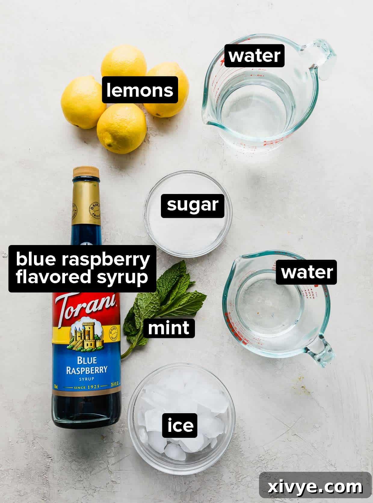 Individual ingredients for blue raspberry lemonade, including fresh lemons, a bottle of blue raspberry syrup, and granulated sugar, arranged on a gray and white marble surface.