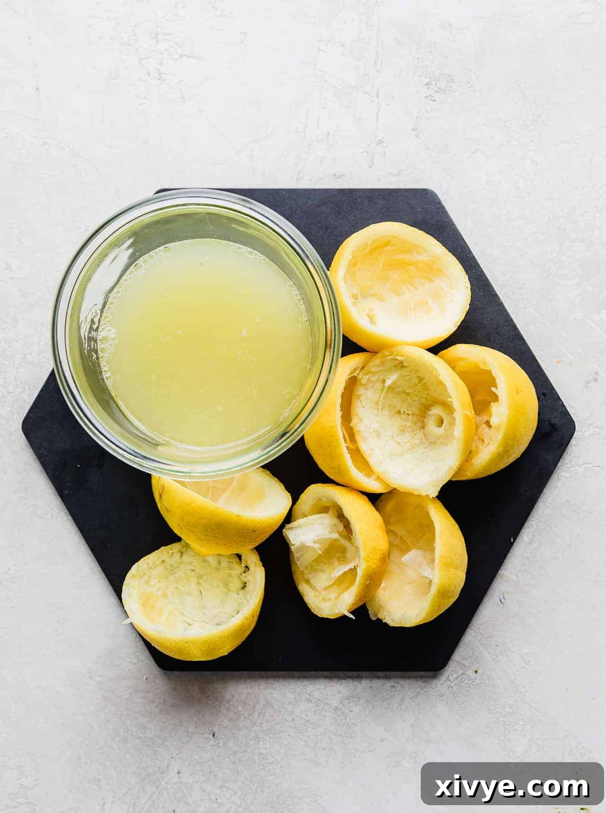 A glass jar filled with freshly squeezed lemon juice, surrounded by squeezed lemon halves and a black hexagon plate, showcasing the key ingredient for lemonade.