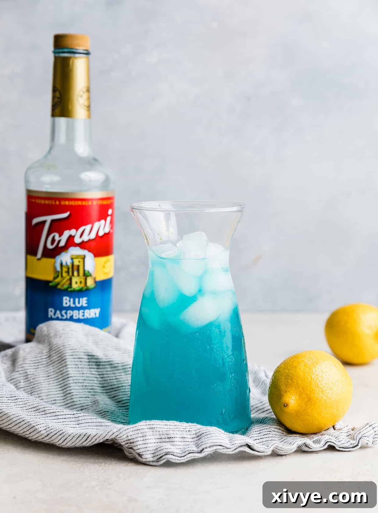 A large glass jar of blue raspberry lemonade standing on a countertop, with a bottle of Torani blue raspberry syrup blurred in the background, showcasing the key flavoring agent.
