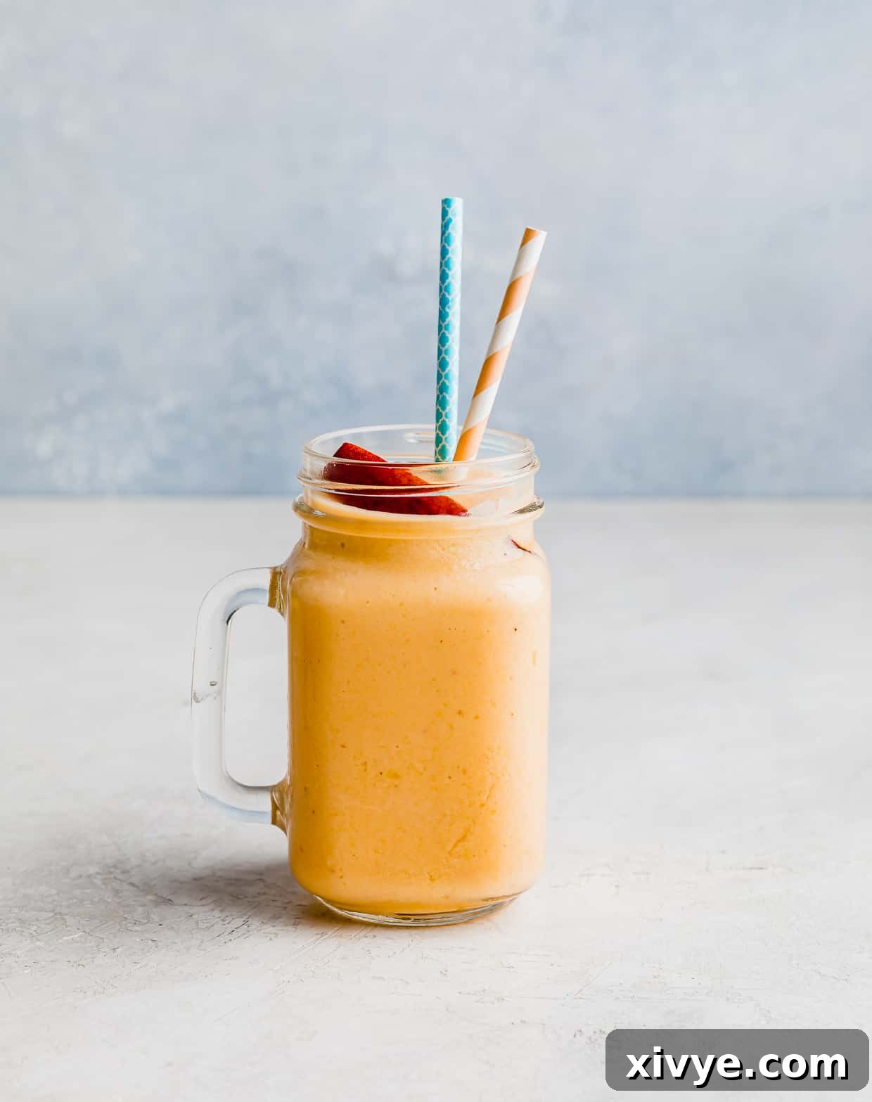 A glass cup filled with an orange colored peaches and banana smoothie against a light blue background.