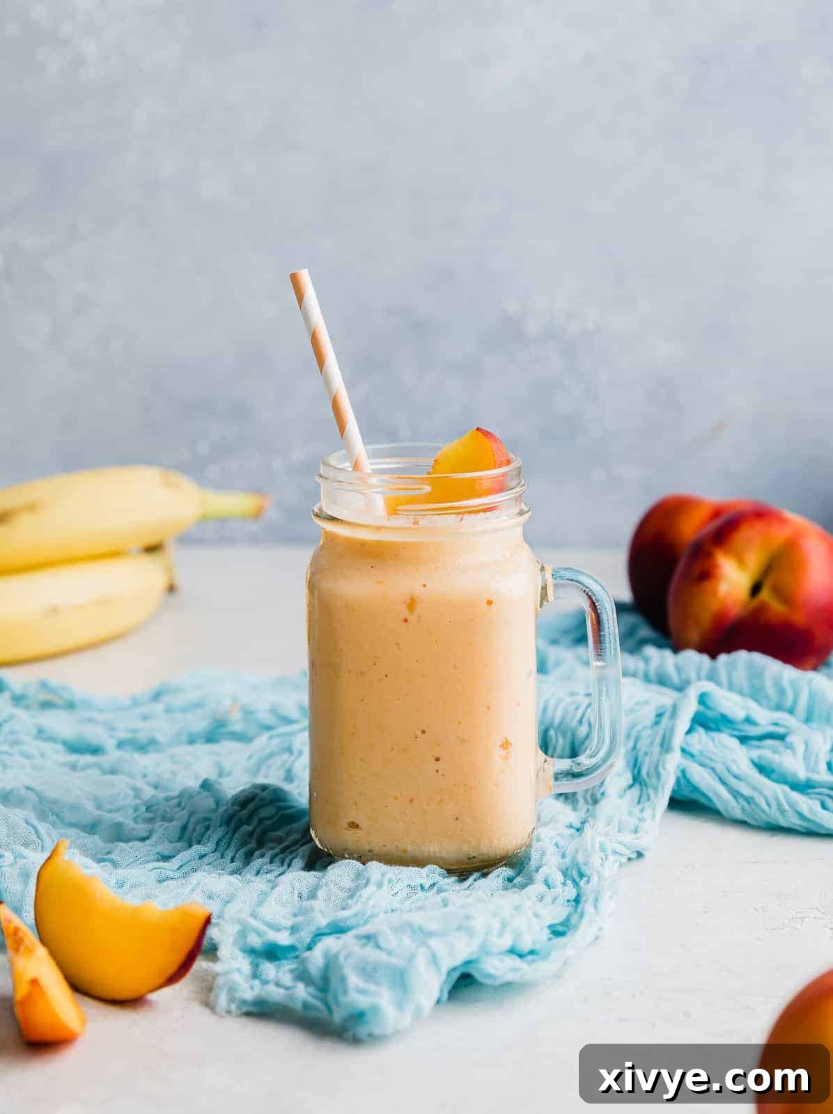 A banana peach smoothie in a glass cup with fresh peaches and bananas in the background.