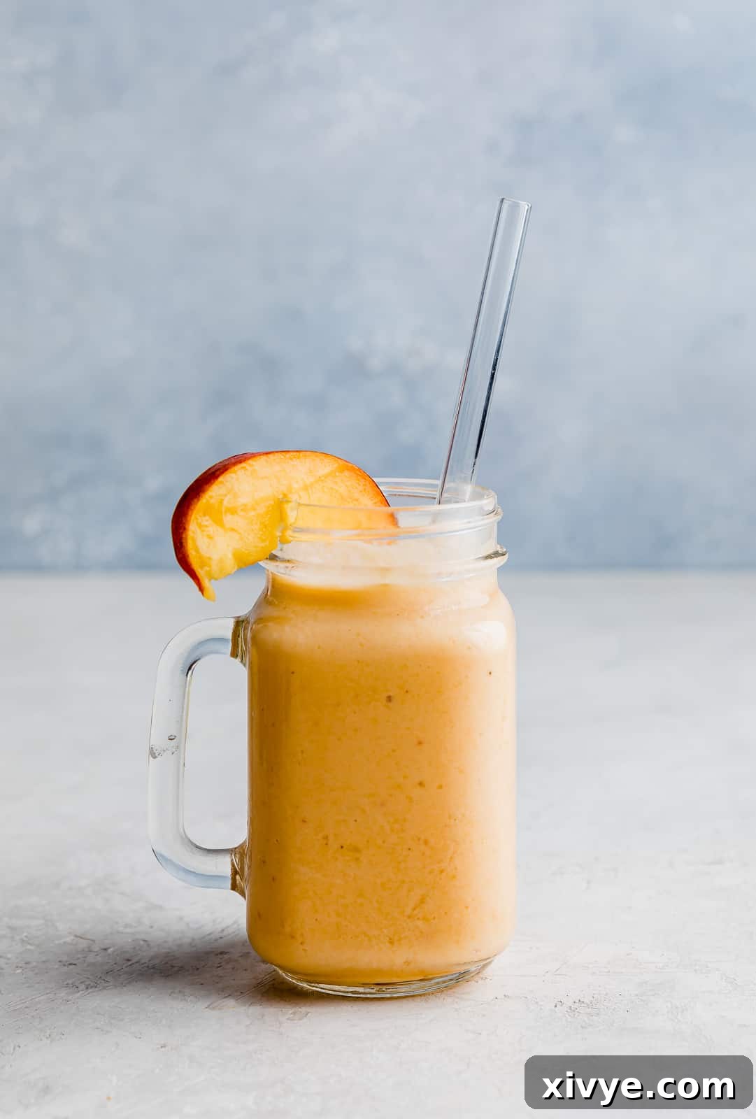 A banana peach smoothie in a glass cup with a fresh peach slice on the rim.