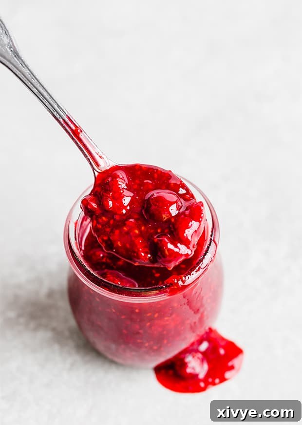 A glass jar on a white background, overflowing with raspberry sauce.