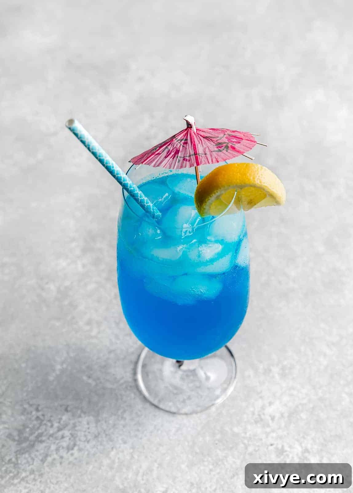 Tropical Azure Cooler 3 A blue lagoon mocktail drink on a gray background with a pink umbrella and lemon wedge on the edge of the cup, evoking a tropical vibe.
