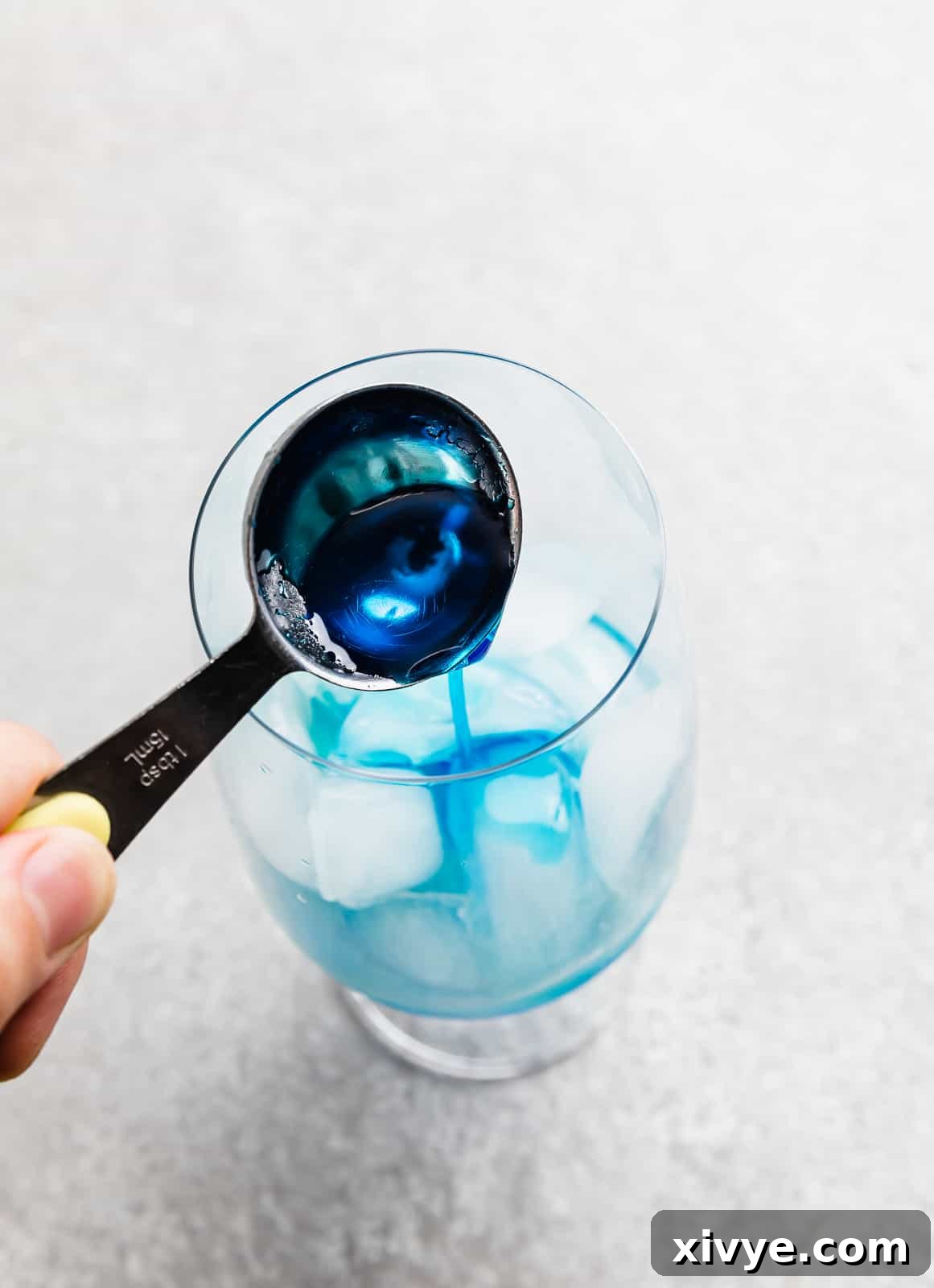 Tropical Azure Cooler 5 A tablespoon full of blue curaçao syrup being poured into a glass cup filled with ice, creating a beautiful swirl.
