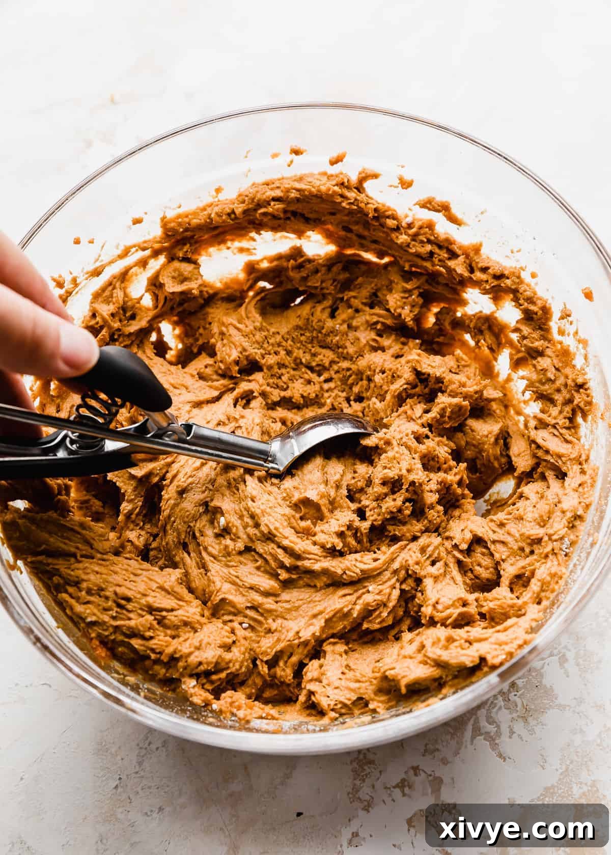 A cookie scooper scooping out orange pumpkin muffin batter from a bowl, ready for baking.