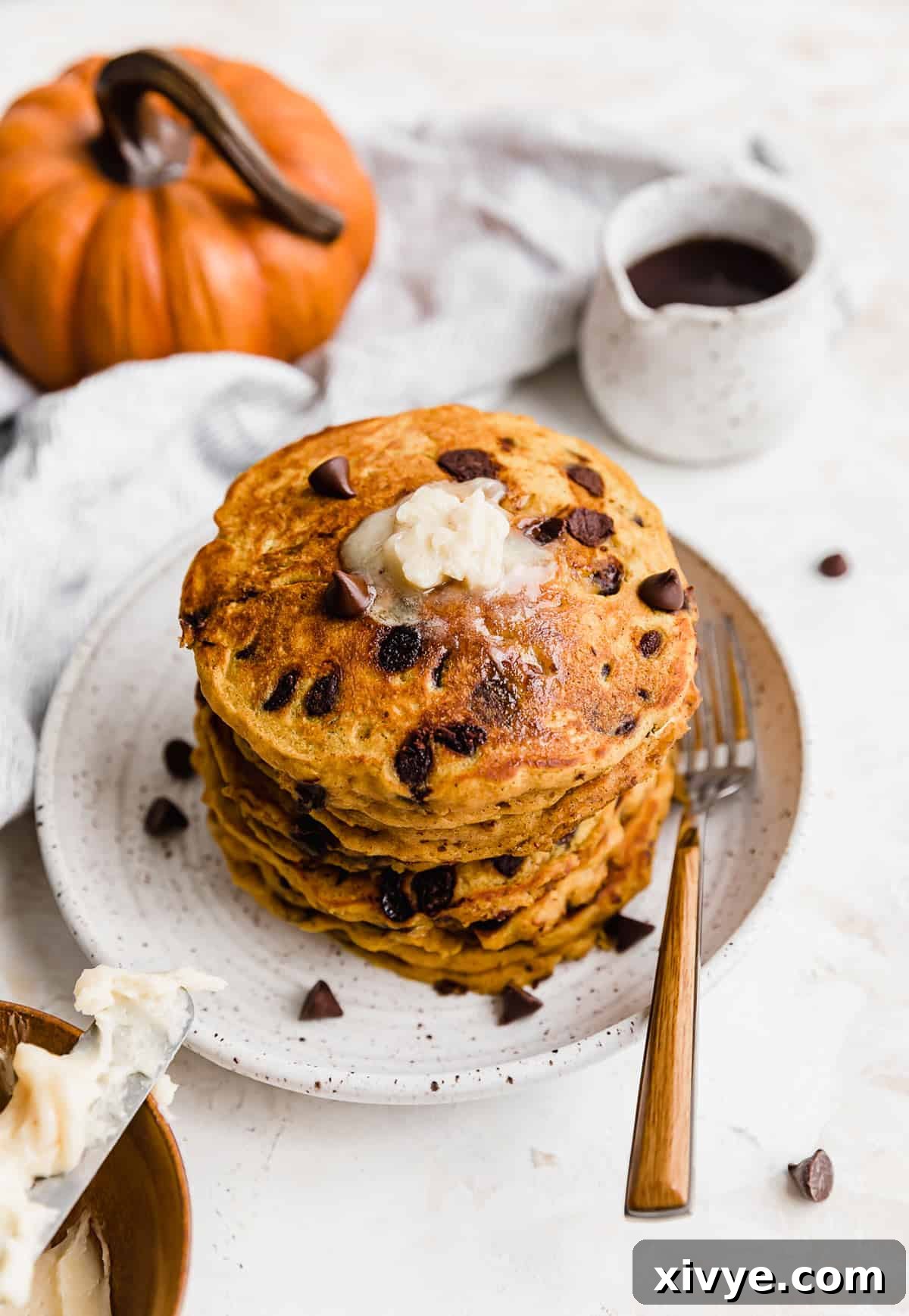A stack of golden brown Pumpkin Chocolate Chip Pancakes on a pristine white plate, drizzled with maple syrup and garnished with a dusting of powdered sugar. The melted chocolate chips are visible, adding to its appetizing appeal.