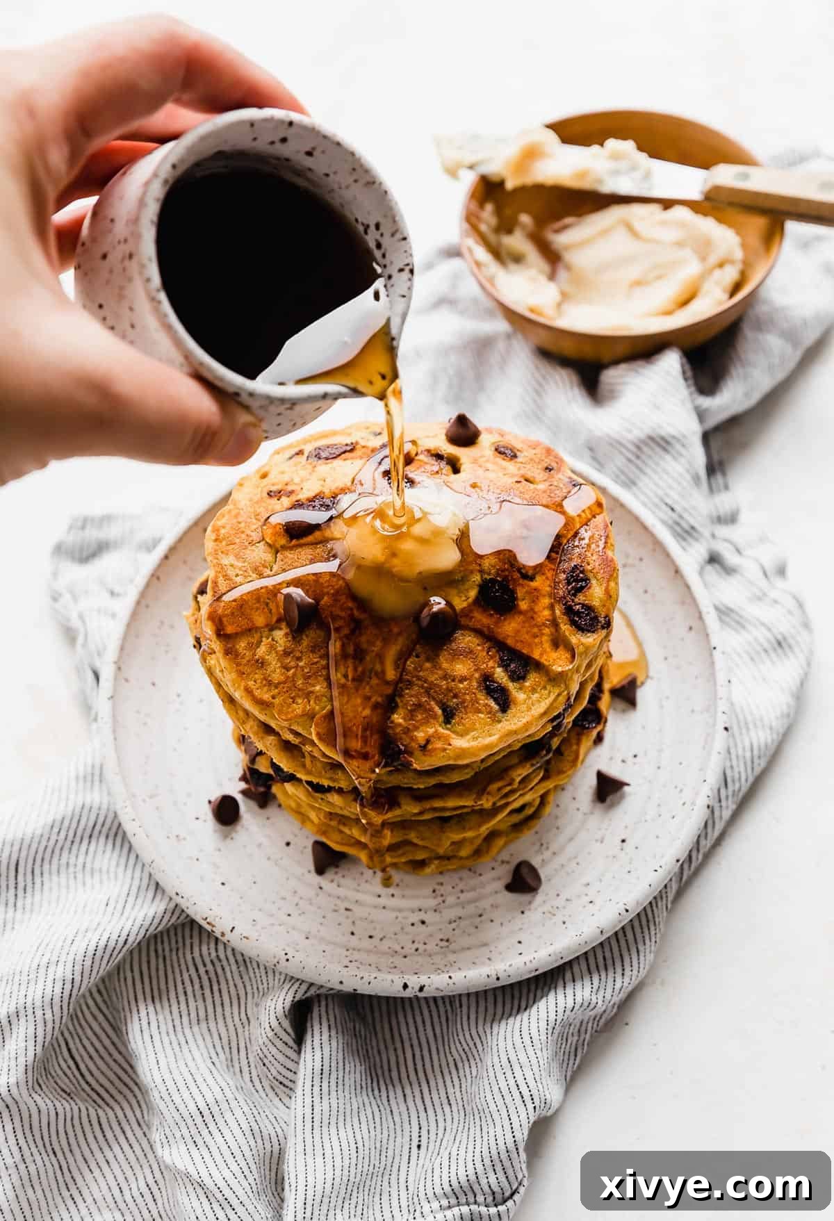 Golden maple syrup being generously poured over a delectable stack of Fluffy Pumpkin Chocolate Chip Pancakes, creating a mouth-watering cascade on a white plate, highlighting their perfect texture.