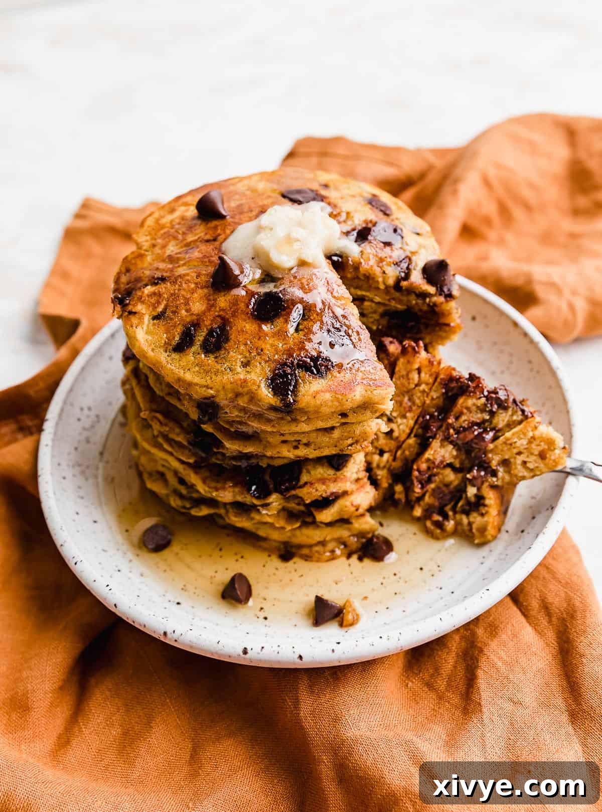 A delectable stack of Pumpkin Chocolate Chip Pancakes on a white plate, with a perfect slice cut out, revealing the moist interior and melted chocolate chips within, ready to be enjoyed.