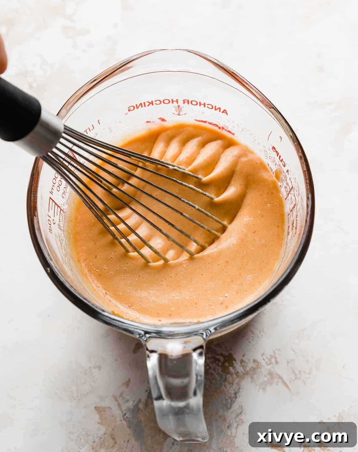 A whisk actively blending an orange-hued liquid mixture in a glass measuring cup, combining pumpkin puree, buttermilk, melted butter, egg yolks, and vanilla for the wet pancake batter.