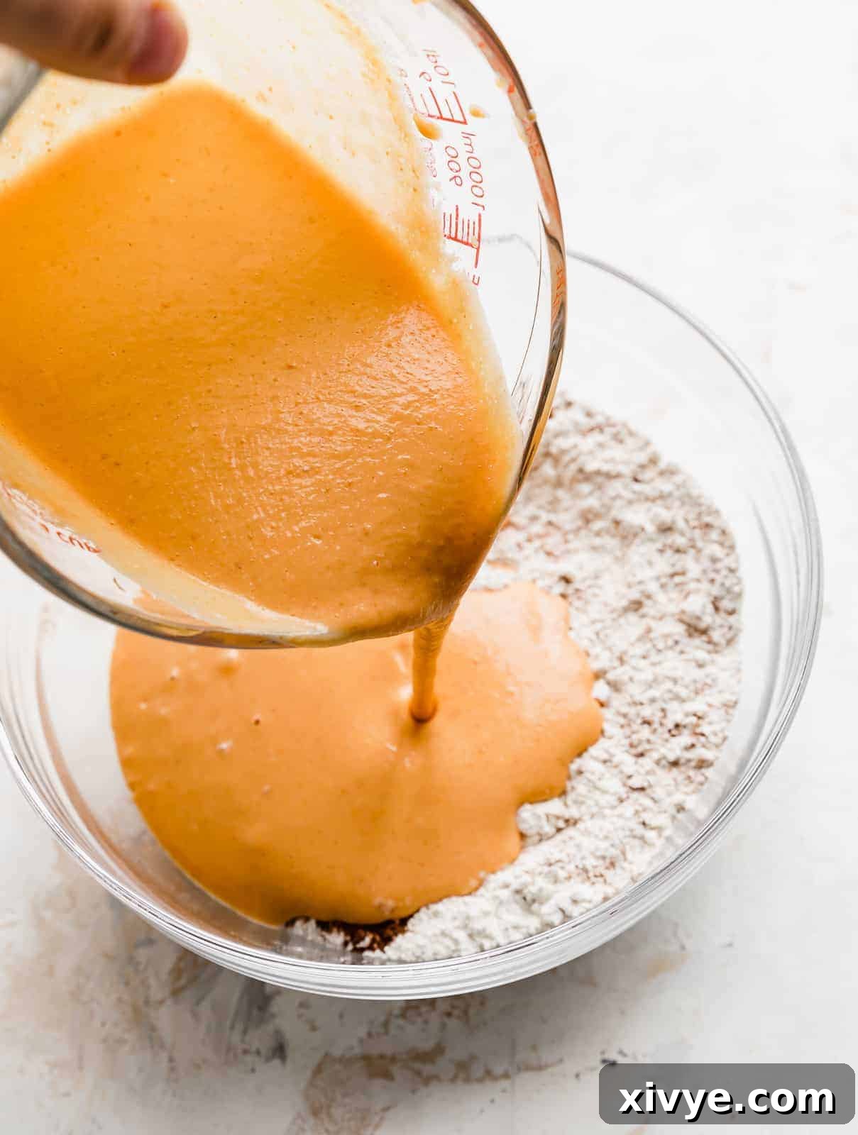 A smooth, orange liquid mixture, containing pumpkin puree and other wet ingredients, being carefully poured into a large bowl filled with dry flour ingredients for pumpkin pancakes.
