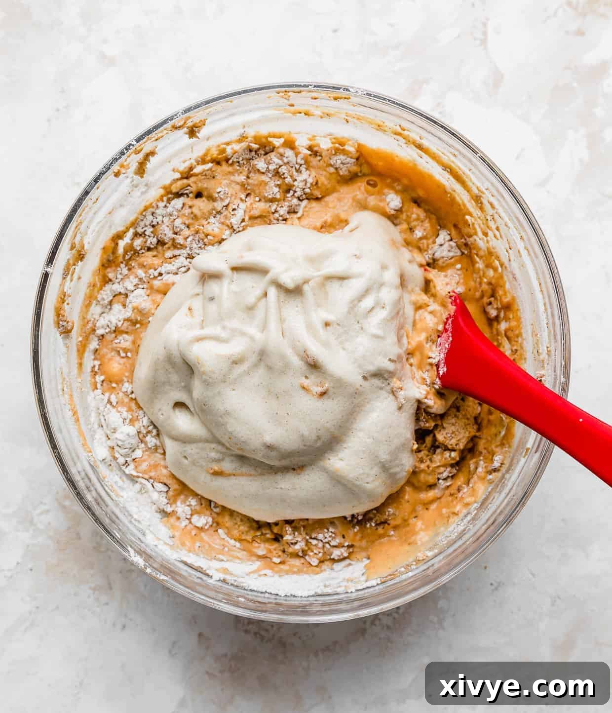 A dollop of fluffy whipped egg whites resting on top of the orange pumpkin pancake batter, ready to be gently folded in for an airy texture.