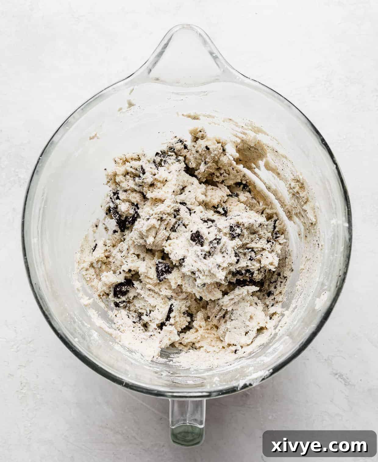 A glass bowl containing a rich, white cookie dough batter, generously studded with dark, chunky Oreo pieces stirred into the mix.
