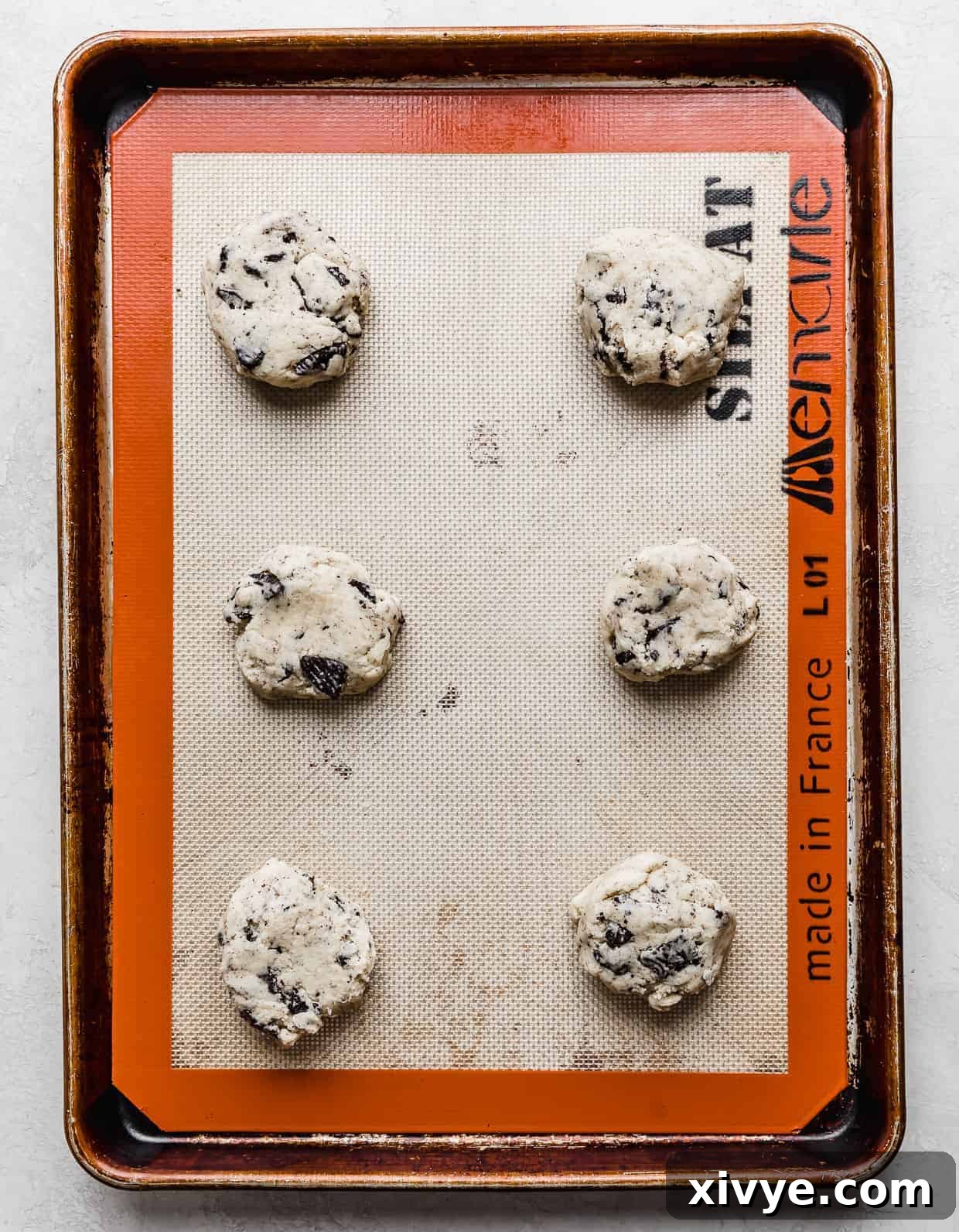 Six perfectly portioned cookies and cream cookie dough balls, lightly pressed into disc shapes, arranged on a baking sheet, ready for the oven.