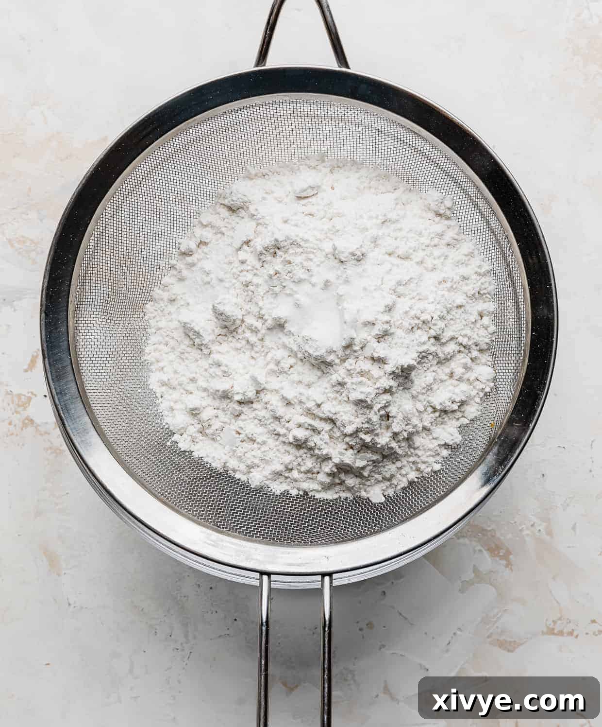 A metal sieve expertly sifting a mixture of cake flour, superfine sugar, and salt into a mixing bowl for angel food cake preparation.