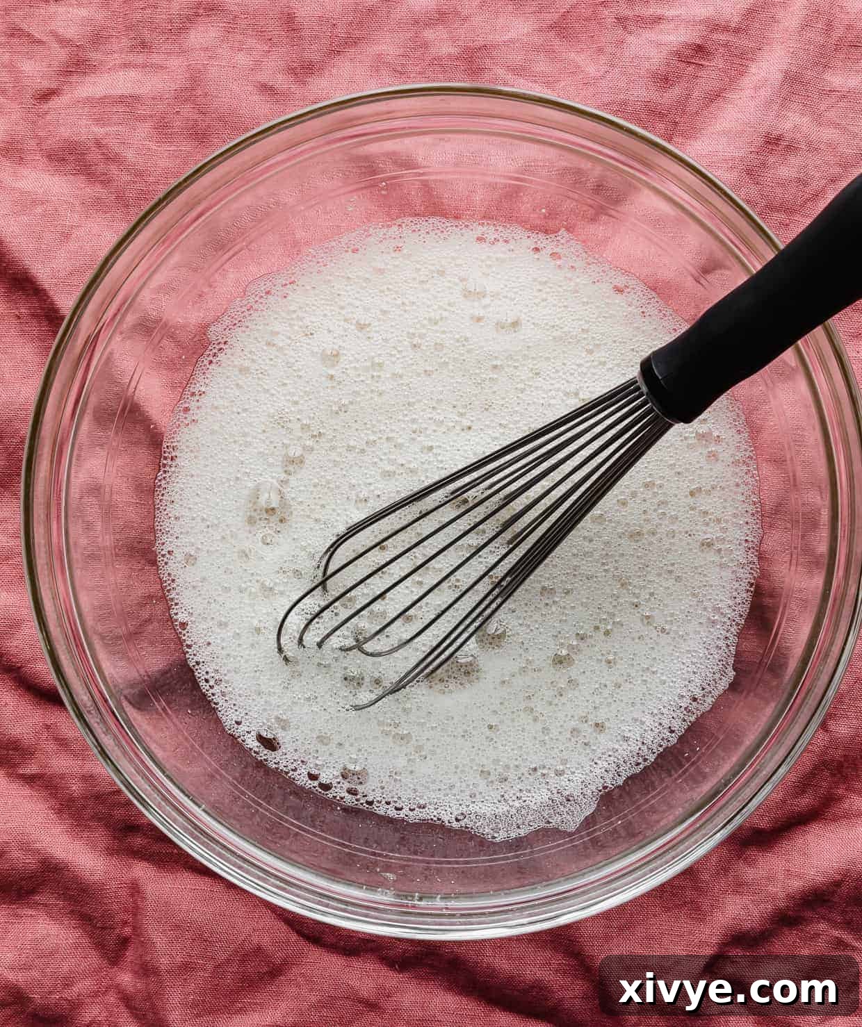 A clear glass bowl showcasing frothy, whisked egg whites, set against a vibrant red linen napkin.