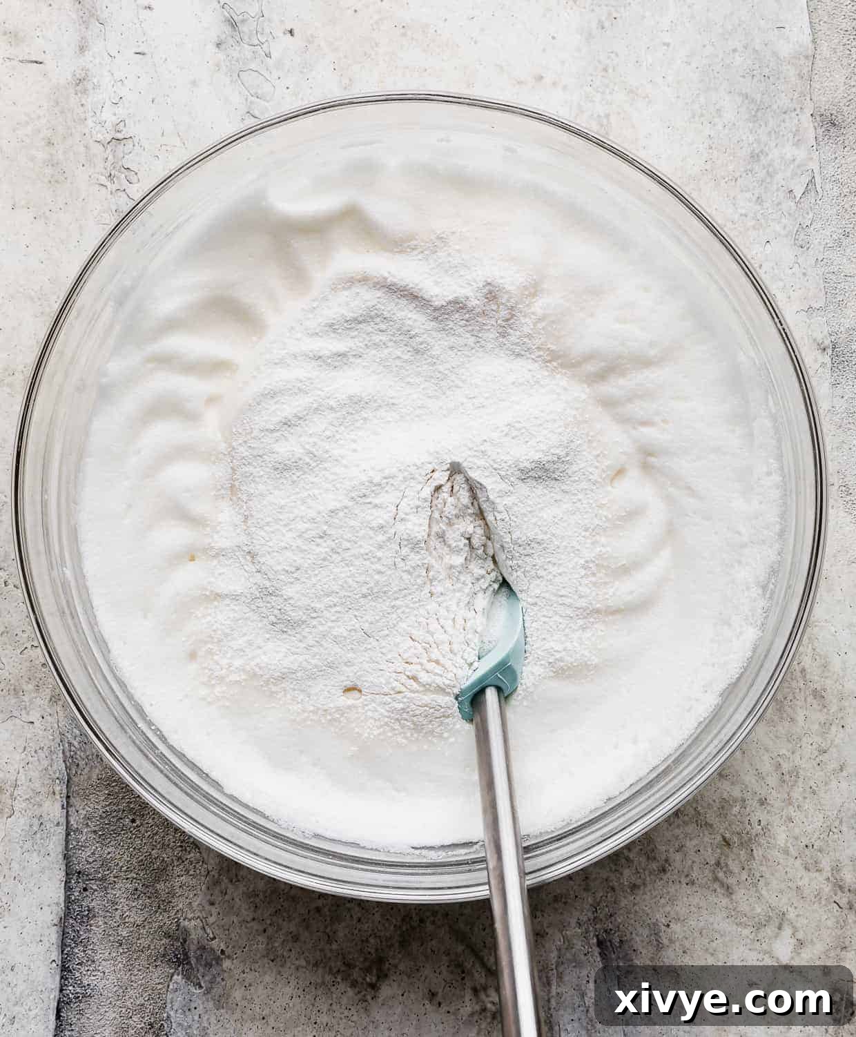 A glass bowl containing beautifully whipped egg whites, with a fine dusting of flour mixture spread delicately over their surface.