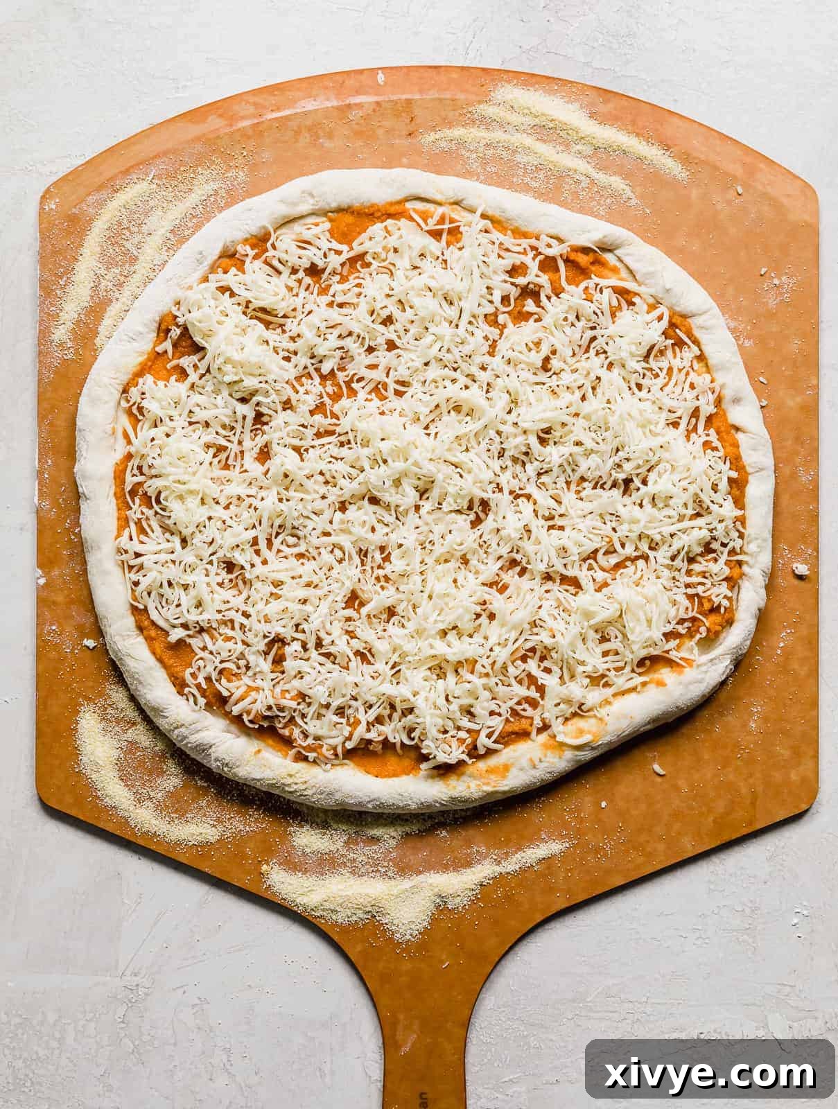 Shredded gouda cheese sprinkled over a pumpkin pizza on a pizza peel.