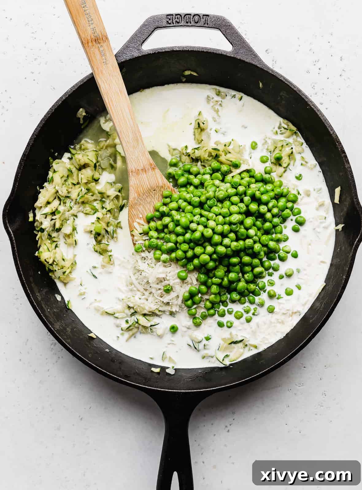 Creamy Zucchini Pasta 8 A black skillet filled with a vibrant mixture of frozen peas, shredded zucchini, and a rich white sauce, simmering gently.