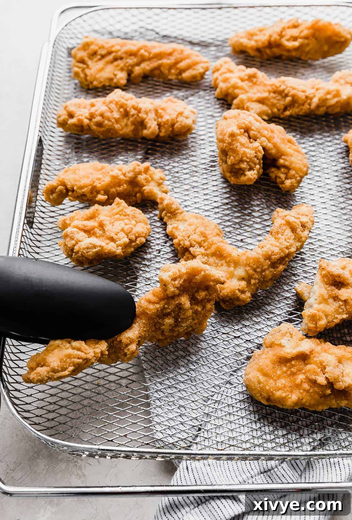 Black kitchen tongs carefully flipping a frozen chicken tender in an air fryer basket, showing the golden side.
