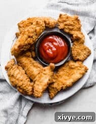Air Fryer Frozen Chicken Tenders on a white plate with a black bowl in the center full of red ketchup.