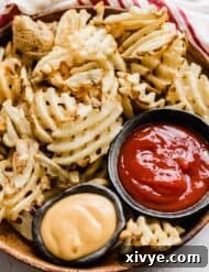 Crispy Waffle Fries from Your Air Fryer 9 Waffle fries on a brown plate with bowls of ketchup and chick fil a sauce beside them.