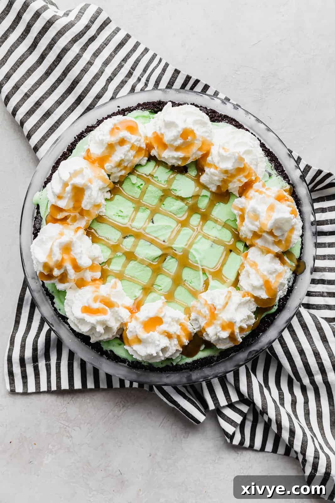 Concrete Crumble 6 A close-up of a completed Asphalt Pie, showcasing luscious caramel drizzled over whipped cream and a sprinkle of crushed Oreos, resting on a black and white striped napkin.