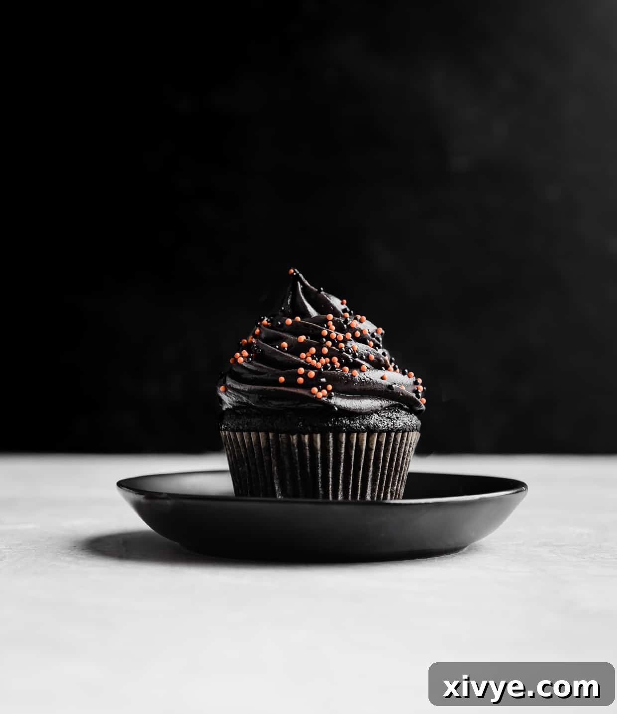 A single Black Velvet Cupcake, beautifully frosted and garnished, resting on a sophisticated black plate. The deep black color of the cake and frosting is prominent, making it an ideal spooky or elegant dessert.