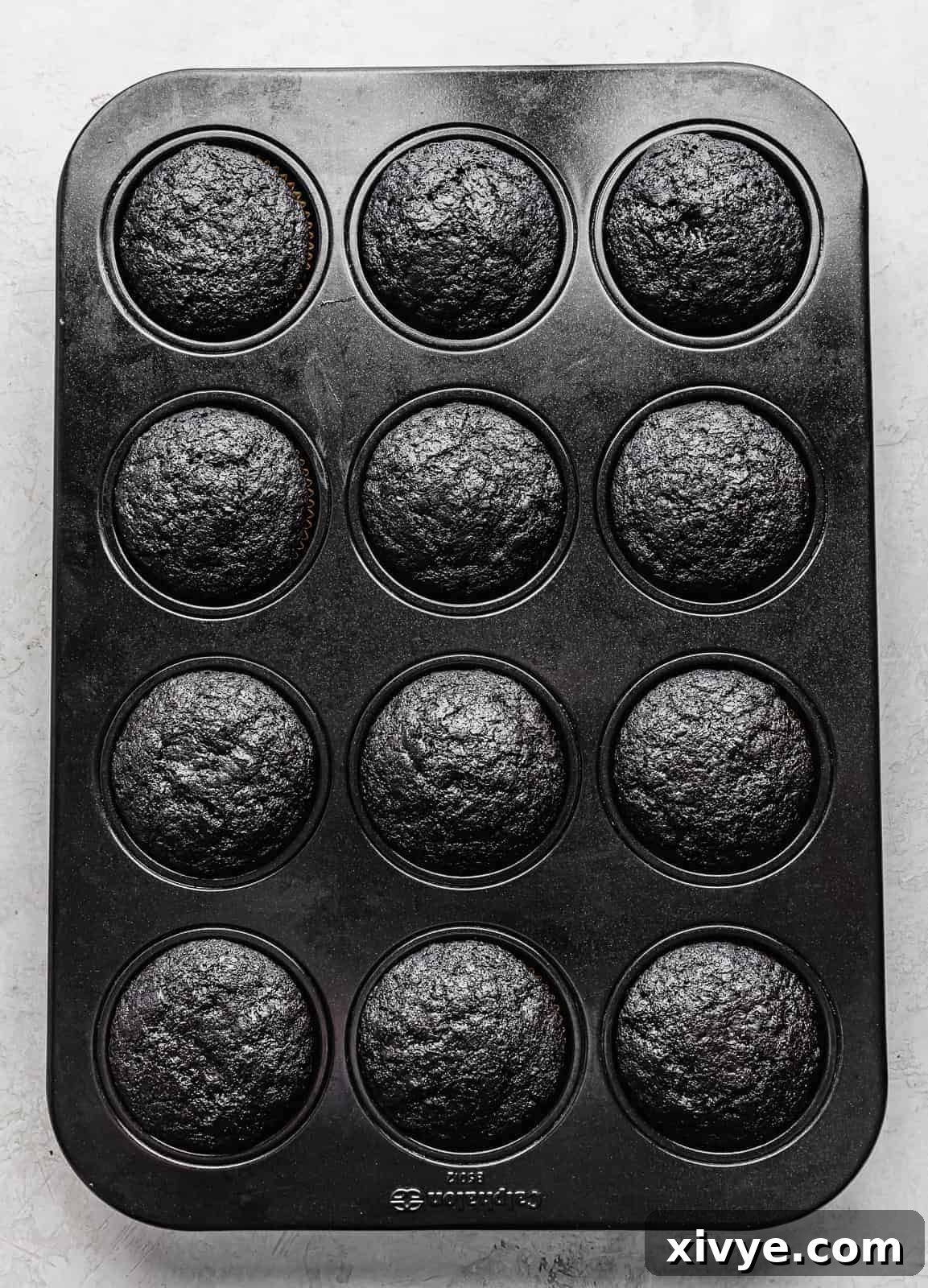 A 12-compartment cupcake pan filled with freshly baked Black Velvet Cupcakes, risen and cooled, showcasing their dark color.
