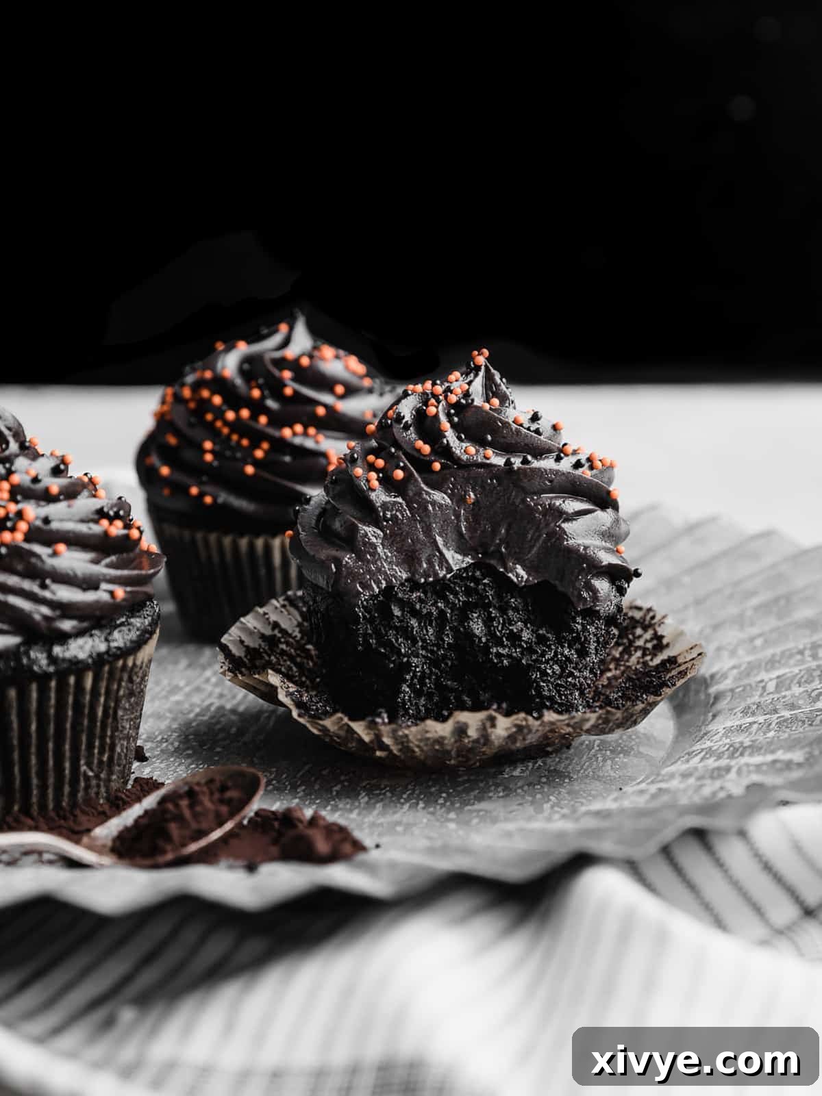 A Black Velvet Cupcake displayed on a gray metal plate, with a bite playfully taken out, revealing the soft, dark interior and fluffy texture against the dark background.