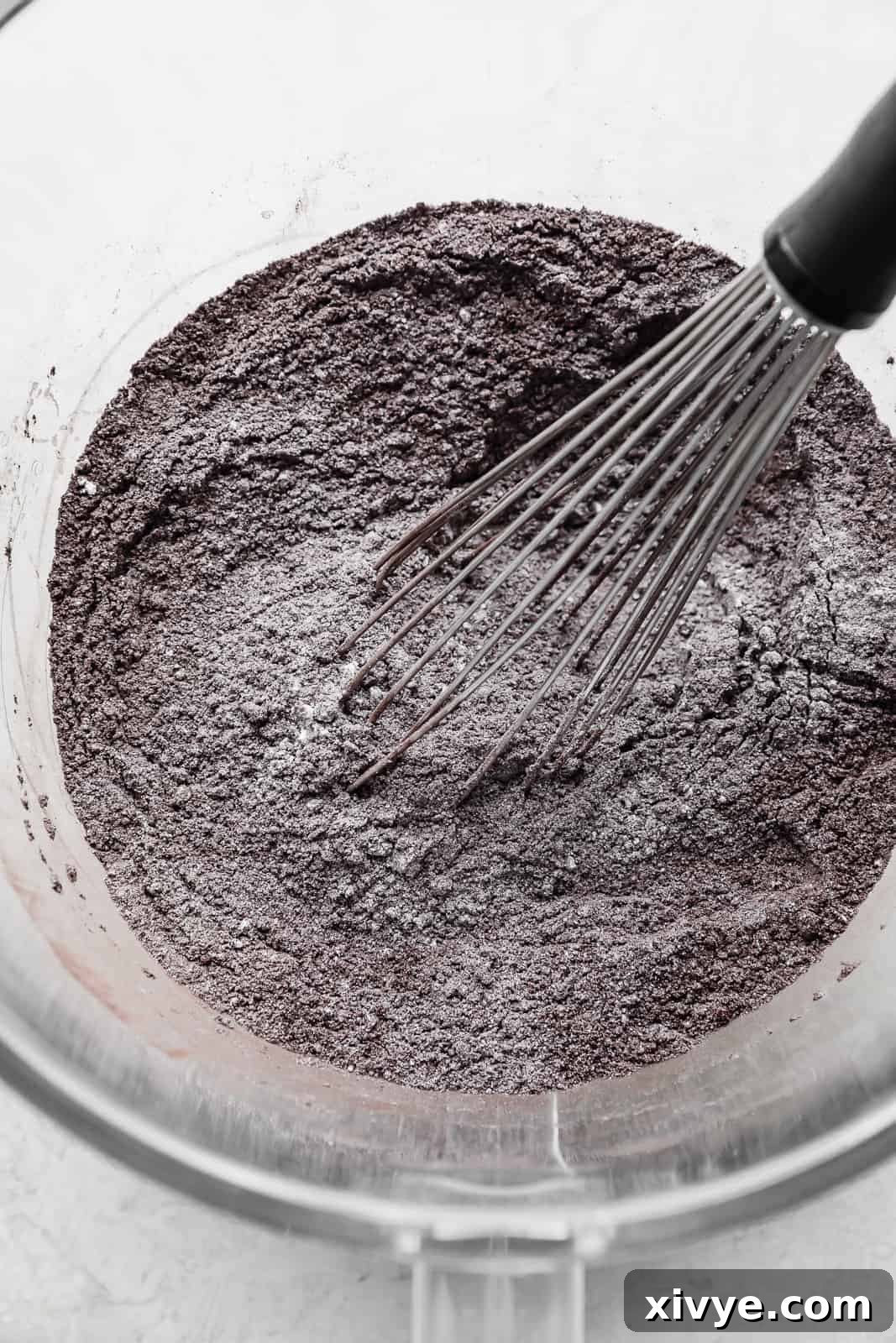 A whisk stirring a light gray colored flour mixture in a clear glass bowl, showcasing the initial blending of the dry ingredients for the Black Velvet Cupcakes.
