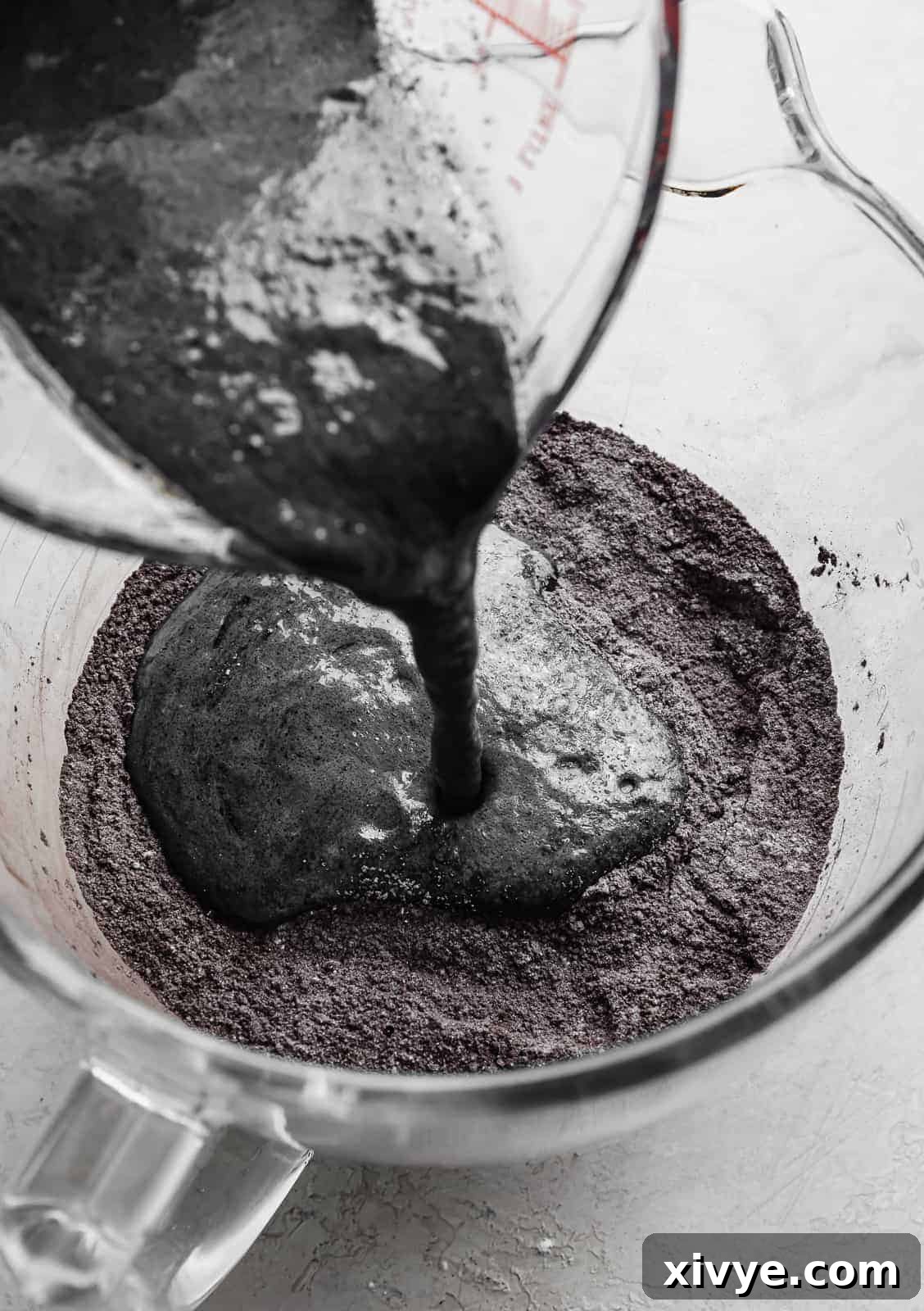The dark liquid wet mixture for Black Velvet Cupcakes being poured into a large glass bowl containing the dry ingredients, initiating the final batter preparation.