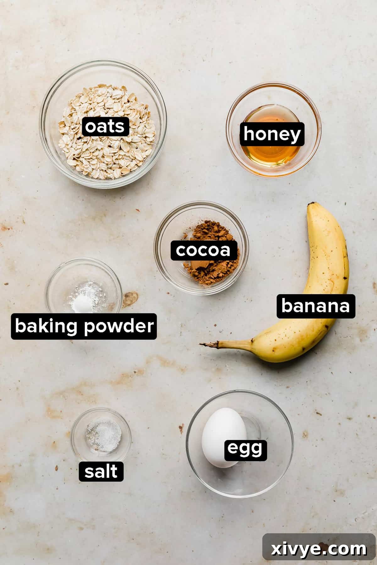 Chocolate Baked Oats ingredients laid out on a cream colored background, including oats, banana, egg, cocoa powder, and honey.