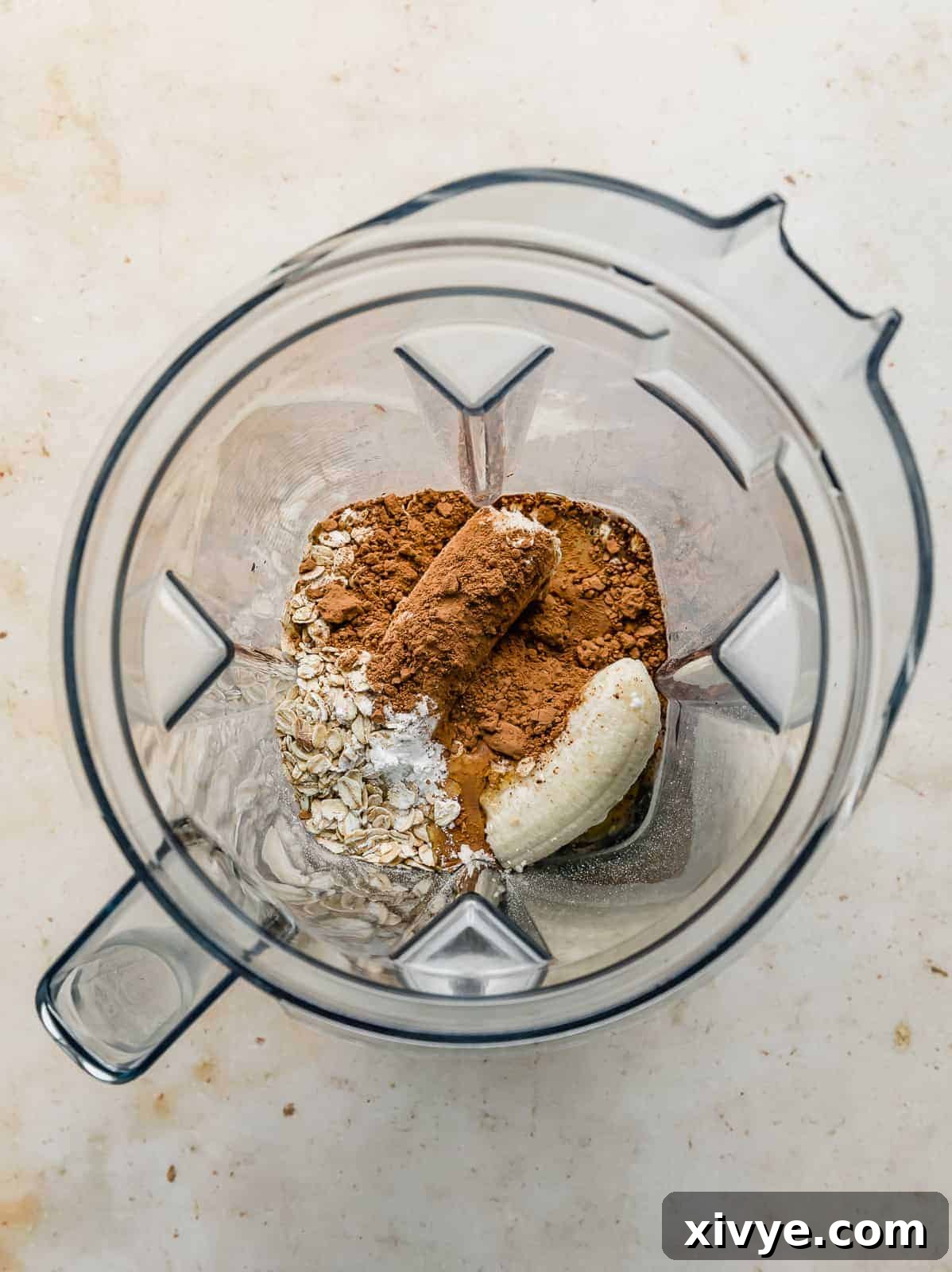 An overhead photo of a blender with a banana, oats, and cocoa unblended.