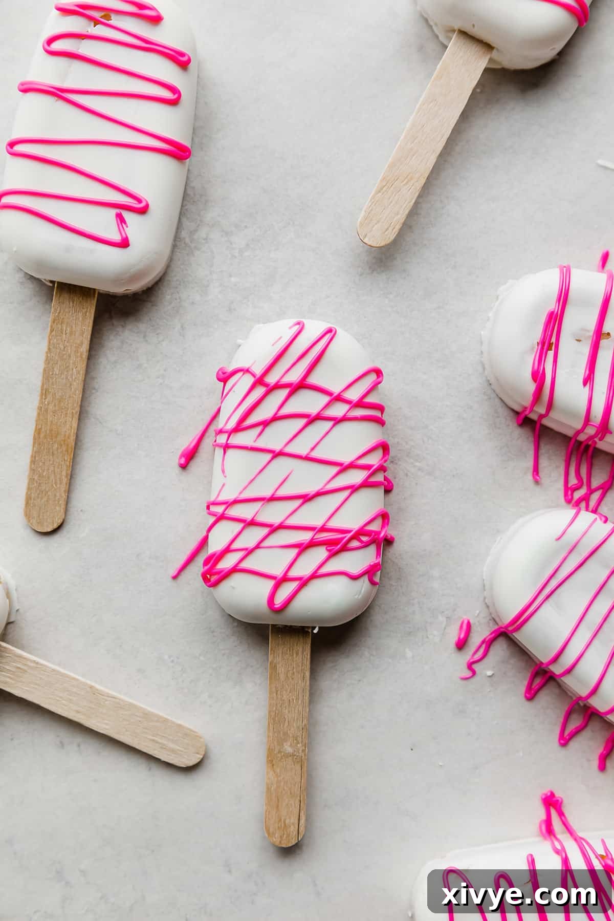 Delightful Cake Lollies 2 Delightful white chocolate-covered Cakesicles, elegantly drizzled with hot pink chocolate, resting on a clean white background.