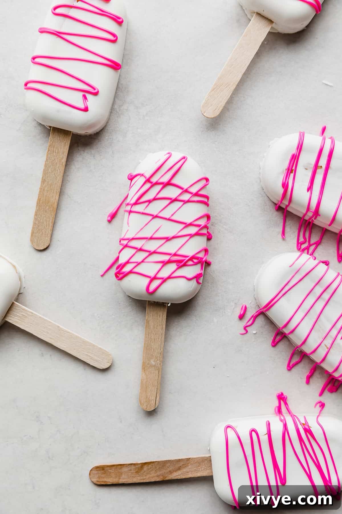 Delightful Cake Lollies 12 An array of white chocolate Cakesicles, each beautifully drizzled with vibrant hot pink chocolate, presented on a clean white background.