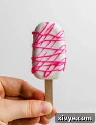 Delightful Cake Lollies 13 A hand holds a white chocolate covered Cakesicle, artfully drizzled with hot pink melted chocolate.