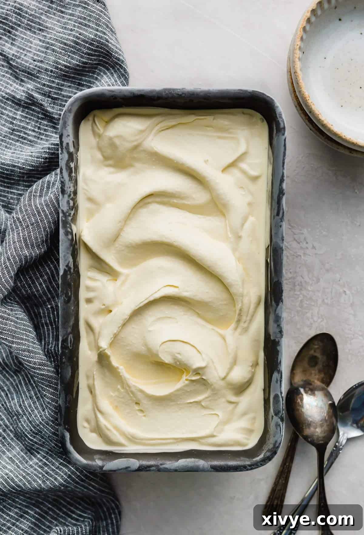 Creamy vanilla ice cream in a dark colored bread pan on a light grey background with spoons next to the pan.