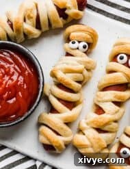 Mummy Hot Dogs on a white plate with a bowl of ketchup to the left.