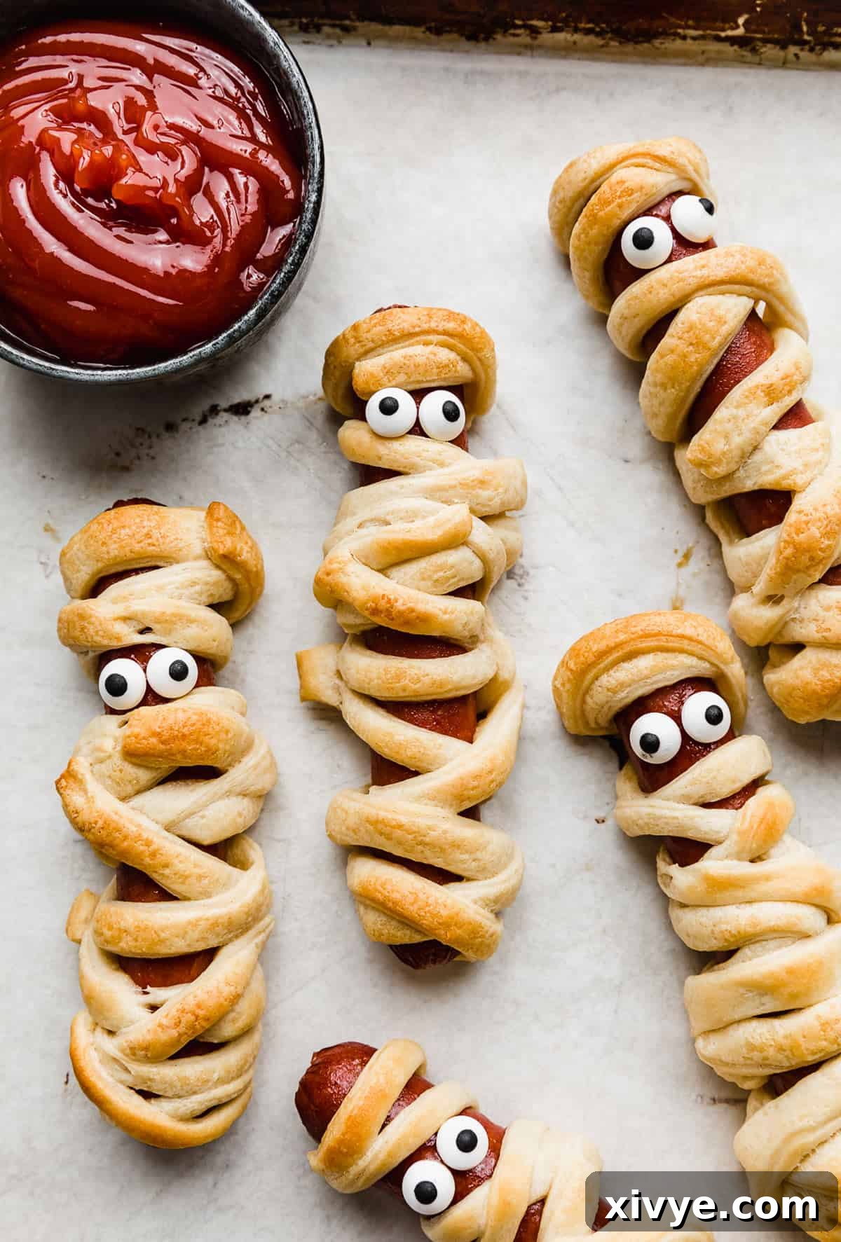 Creepy Canine Wraps 9 Mummy Hot Dogs on a parchment lined baking sheet with candy eyes on the hot dog.